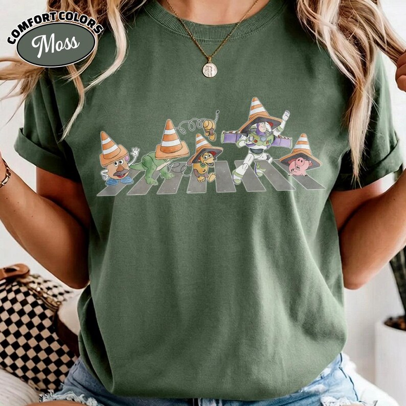 K&ouml;nnte beinhalten: Gr&uuml;nes Comfort Colors T-Shirt mit einer Grafik von Zeichentrickfiguren, die einen Zebrastreifen &uuml;berqueren, jede mit einem Verkehrskegel-Hut. Das Shirt hat den Text "Comfort Colors" und "Moss" in einem kreisf&ouml;rmigen Design.