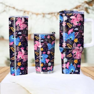 May include: Three insulated tumblers with a repeating pattern of blue and pink cartoon characters, flowers, and leaves on a dark background. The tumblers vary in size and shape, including a tall, slender one, a shorter, wider one, and a mug with a handle.