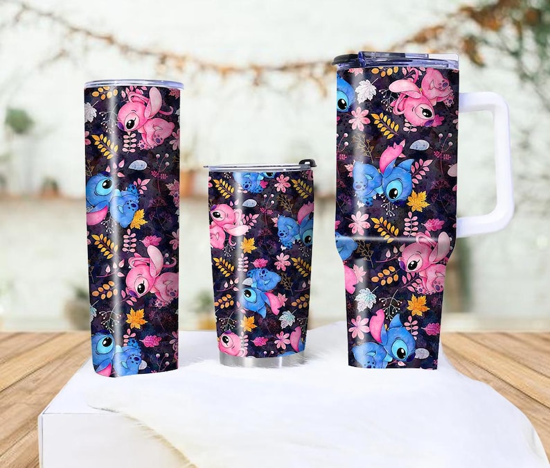 May include: Three insulated tumblers with a repeating pattern of blue and pink cartoon characters, flowers, and leaves on a dark background. The tumblers vary in size and shape, including a tall, slender one, a shorter, wider one, and a mug with a handle.