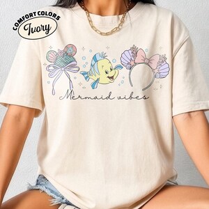 May include: Ivory Comfort Colors t-shirt with a graphic of a cartoon fish and mermaid-themed accessories. The text "Mermaid vibes" is written in a script font. The shirt has a relaxed fit.