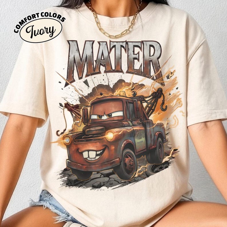 May include: Ivory Comfort Colors t-shirt with a cartoon image of Mater from the film Cars. The design shows Mater with a large grin, set against explosion graphics and the word "MATER" above. The shirt has a crew neck.