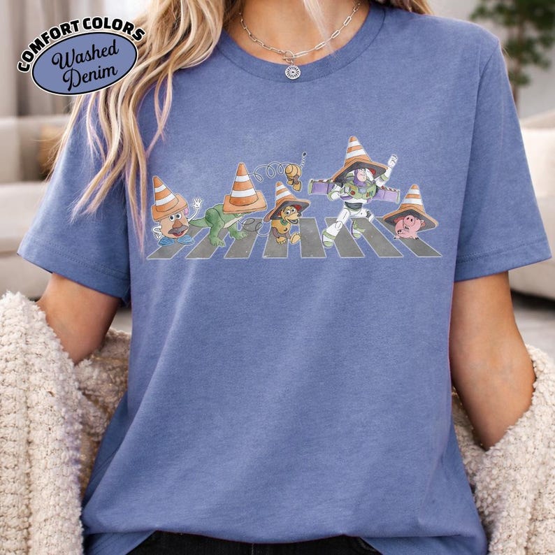 K&ouml;nnte beinhalten: Blaues Comfort Colors Washed Denim T-Shirt mit einer Grafik von Zeichentrickfiguren, die einen Zebrastreifen &uuml;berqueren, jede mit einem Verkehrskegelhut. Der Text "Comfort Colors" und "Washed Denim" befindet sich in einem Kreis oben links.
