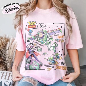 May include: A light pink t-shirt featuring a Toy Story graphic with Buzz Lightyear, Zurg, and green aliens. The design includes the text Toy Story, To infinity & beyond, Galactic Hero, Space Rangger, and Lightyear, set against a space-themed background.