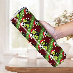 May include: A stainless steel tumbler with a festive design featuring a green character wearing a Santa hat and a black and white character. The tumbler has a red and green diagonal stripe pattern with white snowflakes.