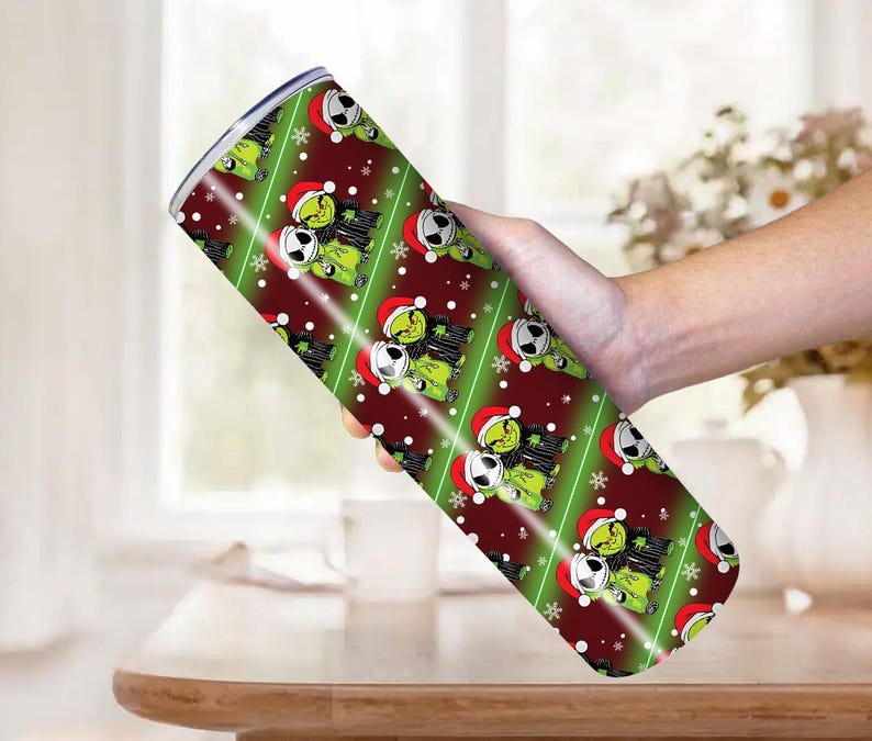 May include: A stainless steel tumbler with a festive design featuring a green character wearing a Santa hat and a black and white character. The tumbler has a red and green diagonal stripe pattern with white snowflakes.