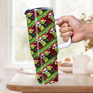 May include: A tall, insulated tumbler with a white handle, featuring a repeating pattern of green Grinch and Jack Skellington characters wearing Santa hats. The background is red with green diagonal stripes and white snowflakes.