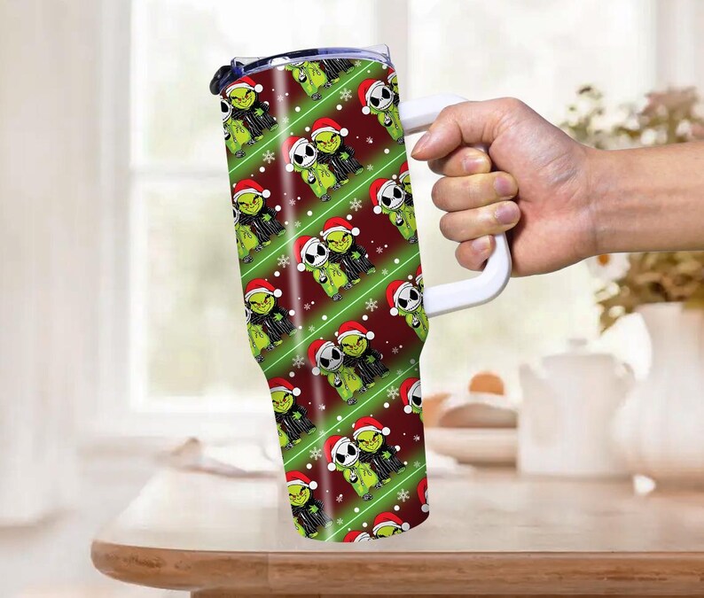 May include: A tall, insulated tumbler with a white handle, featuring a repeating pattern of green Grinch and Jack Skellington characters wearing Santa hats. The background is red with green diagonal stripes and white snowflakes.