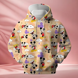 K&ouml;nnte beinhalten: Beiger Kapuzenpullover mit sich wiederholendem Muster von Mickey Mouse-Figuren, Wolken, Sternen und lila Formen. Der Hoodie hat eine wei&szlig;e Kapuze und eine K&auml;ngurutasche. Das Design ist farbenfroh und verspielt.