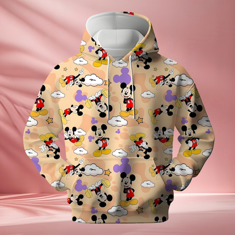 K&ouml;nnte beinhalten: Beiger Kapuzenpullover mit sich wiederholendem Muster von Mickey Mouse-Figuren, Wolken, Sternen und lila Formen. Der Hoodie hat eine wei&szlig;e Kapuze und eine K&auml;ngurutasche. Das Design ist farbenfroh und verspielt.