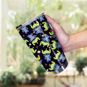 Peut inclure: Gobelet en acier inoxydable avec un motif r&eacute;p&eacute;t&eacute; de personnages de dessins anim&eacute;s bleus et verts sur fond noir. Le motif comprend des &eacute;toiles et d'autres petites formes. Le gobelet a un couvercle noir.