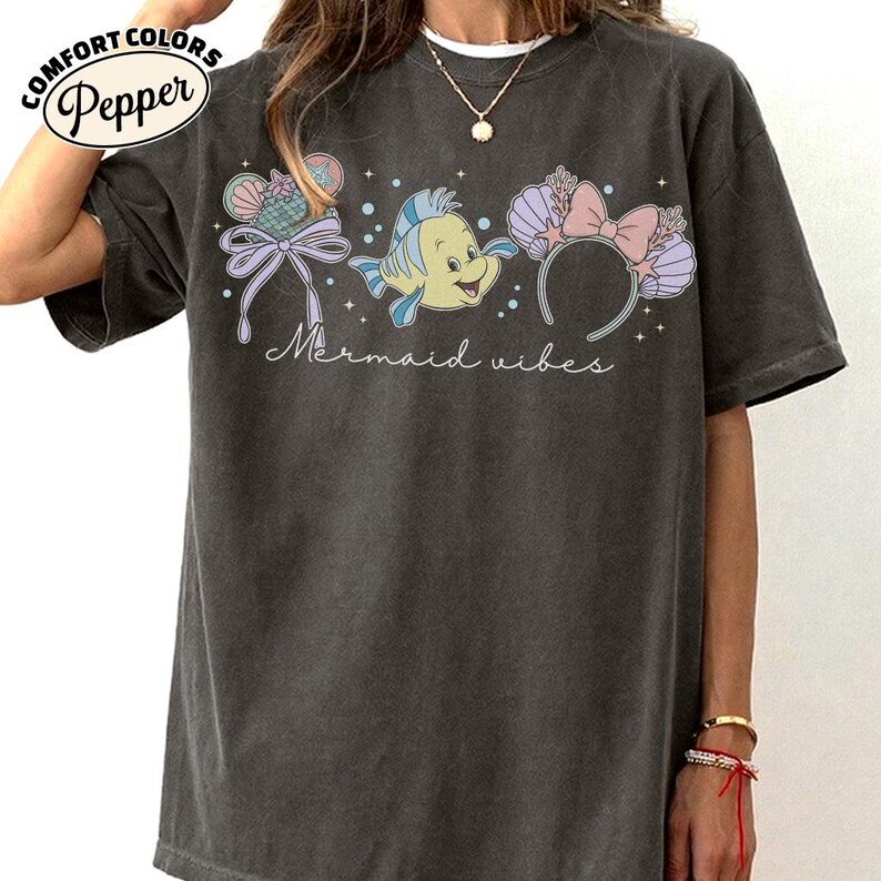 May include: Dark gray Comfort Colors t-shirt with a graphic featuring a cartoon fish, seashell-shaped ears, and a bow. The text "Mermaid vibes" is written in cursive below the graphic. The shirt has a relaxed fit.