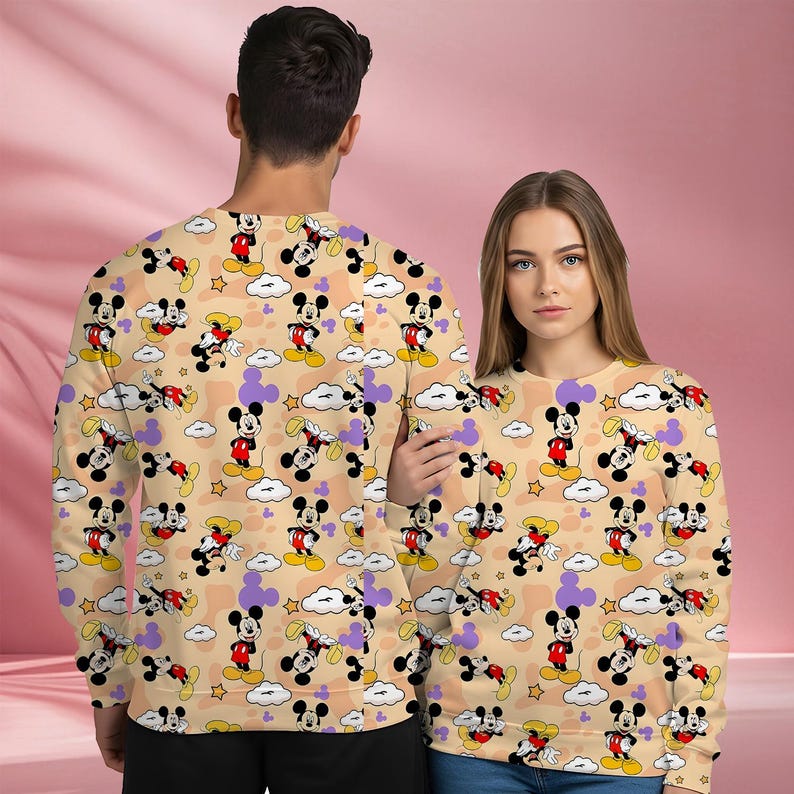 K&ouml;nnte beinhalten: Beige Sweatshirts mit einem sich wiederholenden Muster aus Mickey Mouse, Wolken und Sternen. Mickey Mouse wird in verschiedenen Posen gezeigt, mit seiner charakteristischen roten Hose, gelben Schuhen und wei&szlig;en Handschuhen. Die Sweatshirts sind lang&auml;rmlig.