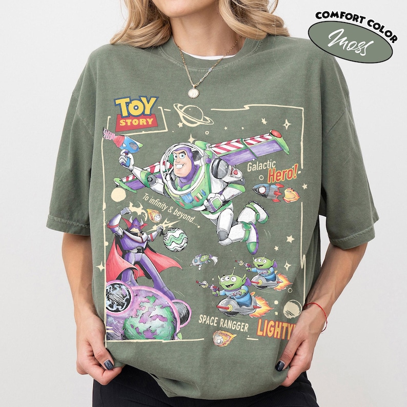 May include: A moss green t-shirt featuring a Toy Story graphic with Buzz Lightyear, Zurg, and aliens. The print includes the text Toy Story, Galactic Hero, To infinity & beyond, Space Rangger, and Lightyear, along with space-themed illustrations.