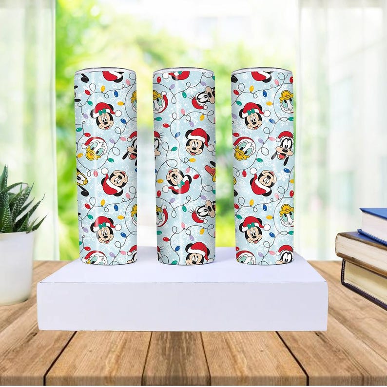 May include: Three cylindrical tumblers with a light blue background featuring Mickey Mouse, Donald Duck, and Goofy in Santa hats and Christmas lights. The tumblers are displayed on a white platform.