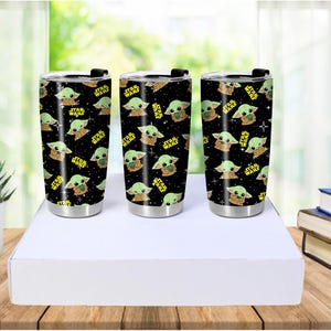 May include: Three stainless steel tumblers with black lids and a repeating pattern of a green alien character and the words "STAR WARS" in yellow. The tumblers are cylindrical and sit on a white surface.