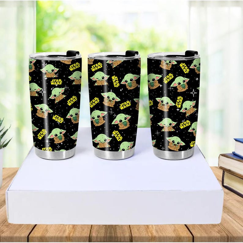 May include: Three stainless steel tumblers with black lids and a repeating pattern of a green alien character and the words "STAR WARS" in yellow. The tumblers are cylindrical and sit on a white surface.