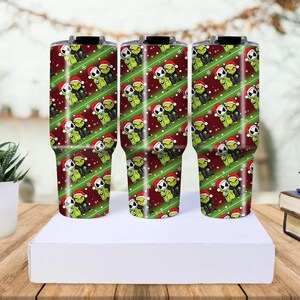 May include: Three stainless steel tumblers with a Christmas-themed design. The tumblers feature a repeating pattern of green characters wearing Santa hats on a red background with green stripes. The tumblers are cylindrical and have black lids.