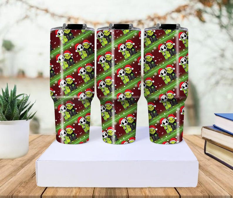 May include: Three stainless steel tumblers with a Christmas-themed design. The tumblers feature a repeating pattern of green characters wearing Santa hats on a red background with green stripes. The tumblers are cylindrical and have black lids.