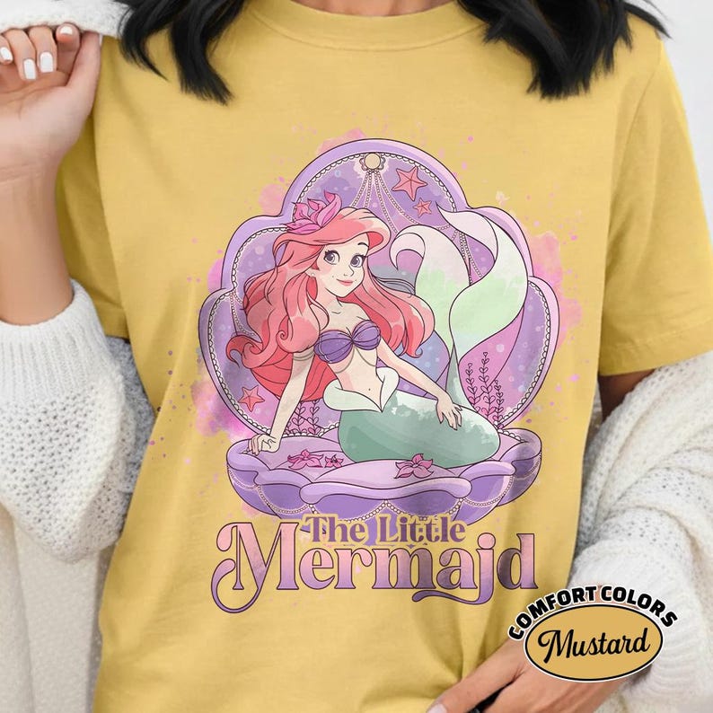 Op de afbeelding: Mosterdkleurig T-shirt met een afbeelding van Ariel uit De Kleine Zeemeermin. Ariel heeft roze haar, een paarse schelpenbeha en een groene staart, zittend in een paarse schelp. De woorden "The Little Mermaid" staan eronder.
