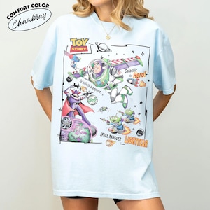 May include: A light blue t-shirt featuring a Toy Story graphic print. The design includes Buzz Lightyear, Zurg, and green aliens in space with text that reads Toy Story, Galactic Hero, To infinity & beyond, Space Rangger, and Lightyear.