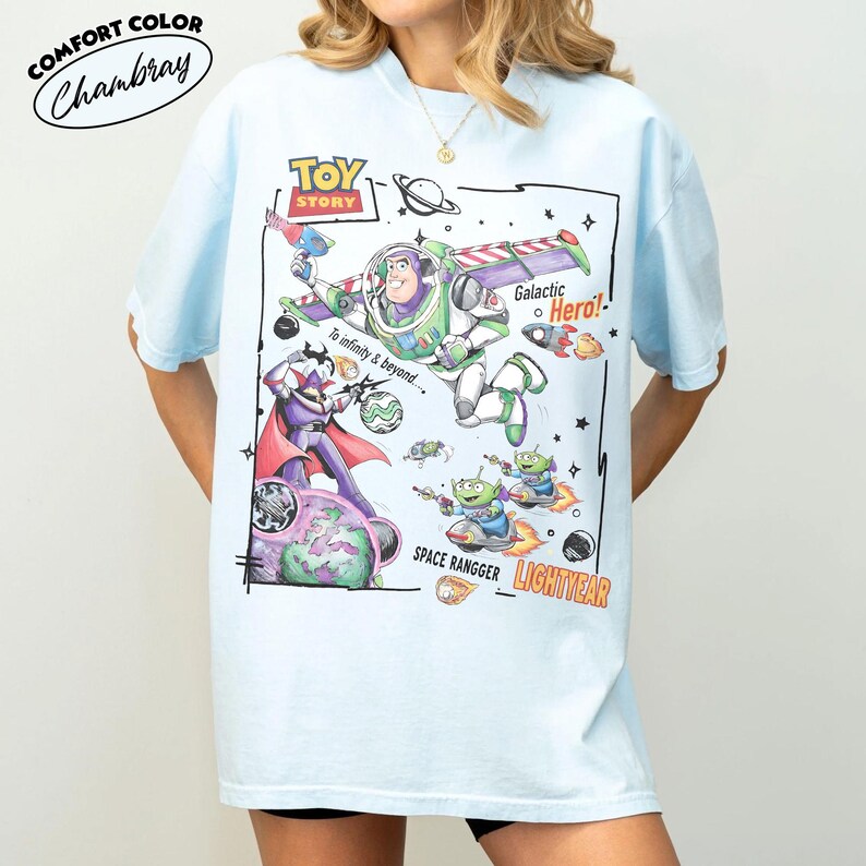 May include: A light blue t-shirt featuring a Toy Story graphic print. The design includes Buzz Lightyear, Zurg, and green aliens in space with text that reads Toy Story, Galactic Hero, To infinity & beyond, Space Rangger, and Lightyear.