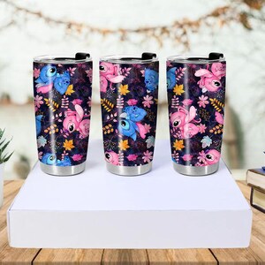 May include: Three stainless steel travel mugs with black lids. Each mug features a dark background with a pattern of blue and pink cartoon characters, flowers, and leaves. The mugs are cylindrical and sit on a white surface.