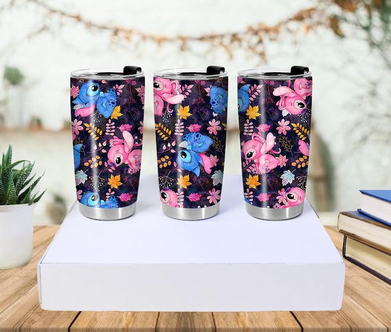 May include: Three stainless steel travel mugs with black lids. Each mug features a dark background with a pattern of blue and pink cartoon characters, flowers, and leaves. The mugs are cylindrical and sit on a white surface.