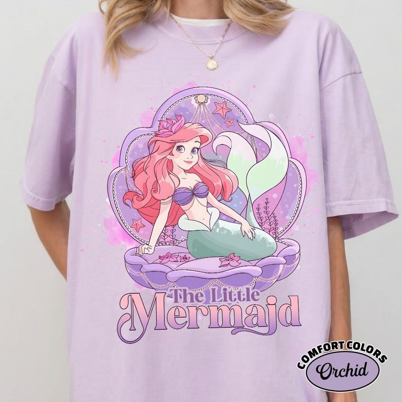 Op de afbeelding: Een lichtpaars T-shirt met een afbeelding van een zeemeermin in een schelp. De zeemeermin heeft roze haar, een paarse top en een groene staart. De tekst "The Little Mermaid" staat eronder. Het shirt heeft de woorden "Comfort Colors" en "Orchid" rechtsonder.