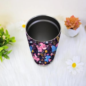 May include: A stainless steel tumbler with a wrap-around design featuring pink and blue cartoon characters, flowers, and leaves on a dark purple background. The tumbler has a silver rim and a dark interior.