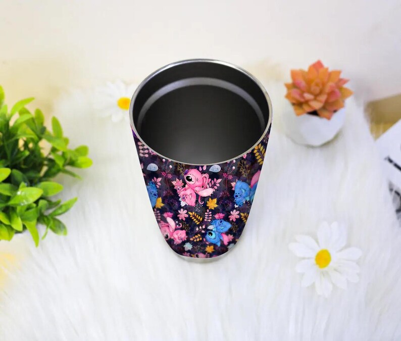 May include: A stainless steel tumbler with a wrap-around design featuring pink and blue cartoon characters, flowers, and leaves on a dark purple background. The tumbler has a silver rim and a dark interior.