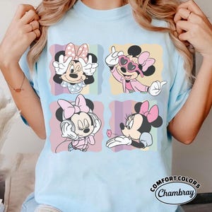 May include: Light blue t-shirt featuring four panels with Minnie Mouse illustrations. Minnie is depicted in various poses, wearing pink bows and outfits. The shirt has the text "Comfort Colors Chambray" at the bottom right.
