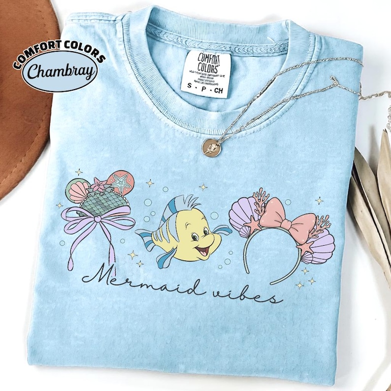 May include: Light blue t-shirt with the words "Mermaid Vibes" and a graphic of a yellow fish and mermaid-themed accessories. The shirt has a crew neck and short sleeves. A silver necklace with a pendant is on the shirt.