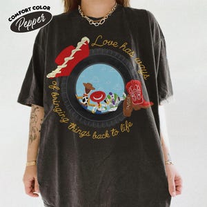 Może przedstawiać: Ciemnoszary t-shirt z kr&oacute;tkim rękawem z grafiką przedstawiającą oponę otaczającą postacie z Toy Story. Napis brzmi Love has ways of bringing things back to life. Na oponie umieszczono czerwony kowbojski kapelusz oraz czerwony kowbojski but.