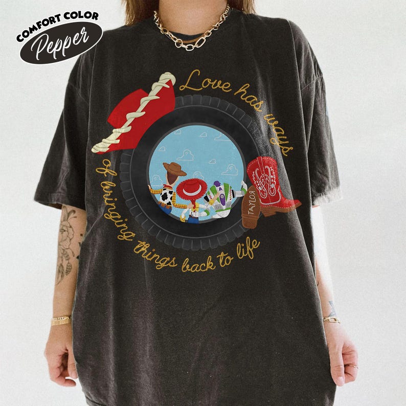 Może przedstawiać: Ciemnoszary t-shirt z kr&oacute;tkim rękawem z grafiką przedstawiającą oponę otaczającą postacie z Toy Story. Napis brzmi Love has ways of bringing things back to life. Na oponie umieszczono czerwony kowbojski kapelusz oraz czerwony kowbojski but.