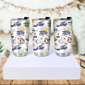 Op de afbeelding: Drie roestvrijstalen reisbekers met zwarte deksels. Elke beker heeft een witte achtergrond met een Star Wars-thema, waaronder de Millennium Falcon, Baby Yoda en andere personages. De bekers worden op een witte doos gepresenteerd.