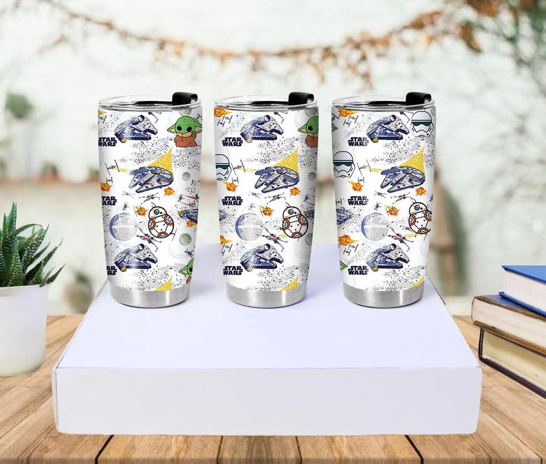 Op de afbeelding: Drie roestvrijstalen reisbekers met zwarte deksels. Elke beker heeft een witte achtergrond met een Star Wars-thema, waaronder de Millennium Falcon, Baby Yoda en andere personages. De bekers worden op een witte doos gepresenteerd.