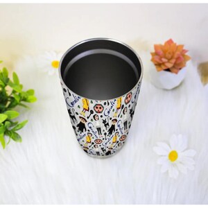 May include: A stainless steel tumbler with a white background and a repeating pattern of cartoon characters, skulls, and the name "Coraline" in blue script. The tumbler has a silver interior and is empty.