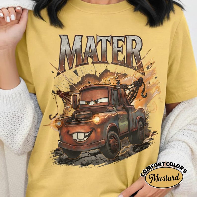 May include: Mustard-coloured t-shirt with a graphic of Mater from the Cars film. The design includes the word "MATER" above the truck, in a distressed style. The shirt has a Comfort Colors label.