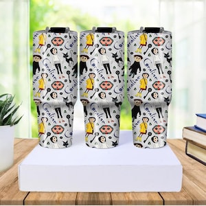 May include: Three white travel mugs with black lids, featuring a repeating pattern of a girl in a yellow raincoat, a black cat, and the word "Coraline". The mugs are on a white box, with a blurred background.