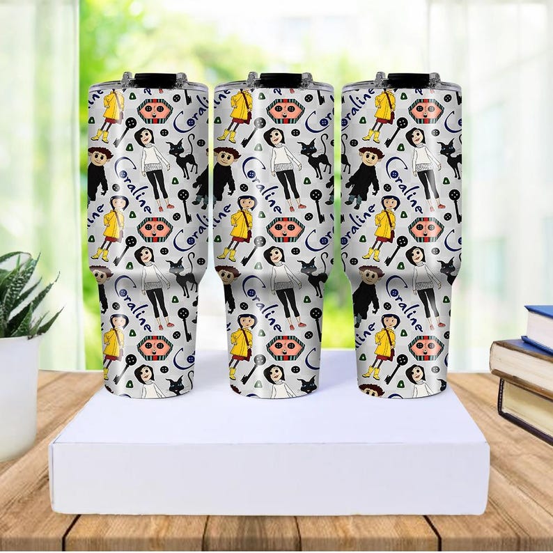 May include: Three white travel mugs with black lids, featuring a repeating pattern of a girl in a yellow raincoat, a black cat, and the word "Coraline". The mugs are on a white box, with a blurred background.