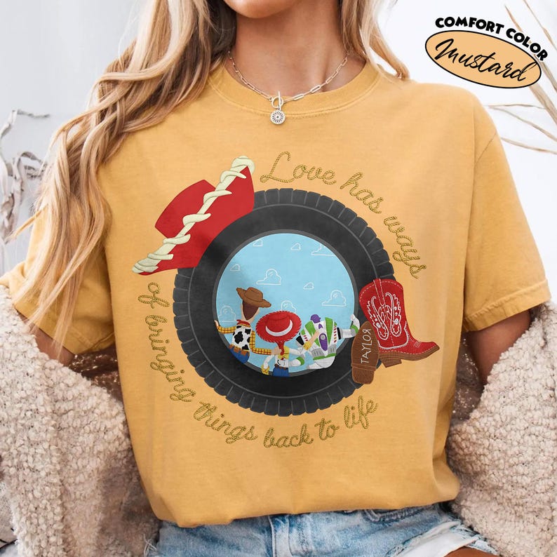 Może przedstawiać: Musztardowy t-shirt z grafiką czarnej opony otaczającej scenę z niebieskim niebem i trzema postaciami z kresk&oacute;wek. Czerwony kowbojski kapelusz leży na oponie, a czerwony kowbojski but znajduje się po prawej stronie. Napis: Love has ways of bringing things back to life.
