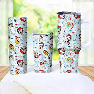 May include: Three insulated tumblers with a light blue background featuring a repeating pattern of Mickey Mouse, Donald Duck, and Goofy in Santa hats, along with Christmas lights. The tumblers vary in size and shape, with one having a handle.