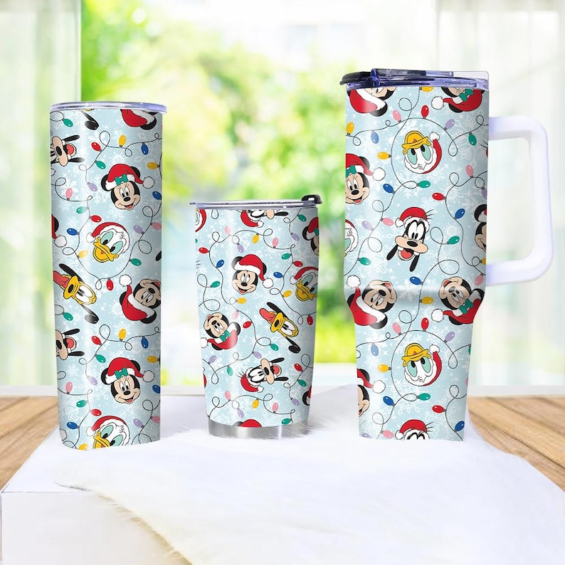 May include: Three insulated tumblers with a light blue background featuring a repeating pattern of Mickey Mouse, Donald Duck, and Goofy in Santa hats, along with Christmas lights. The tumblers vary in size and shape, with one having a handle.