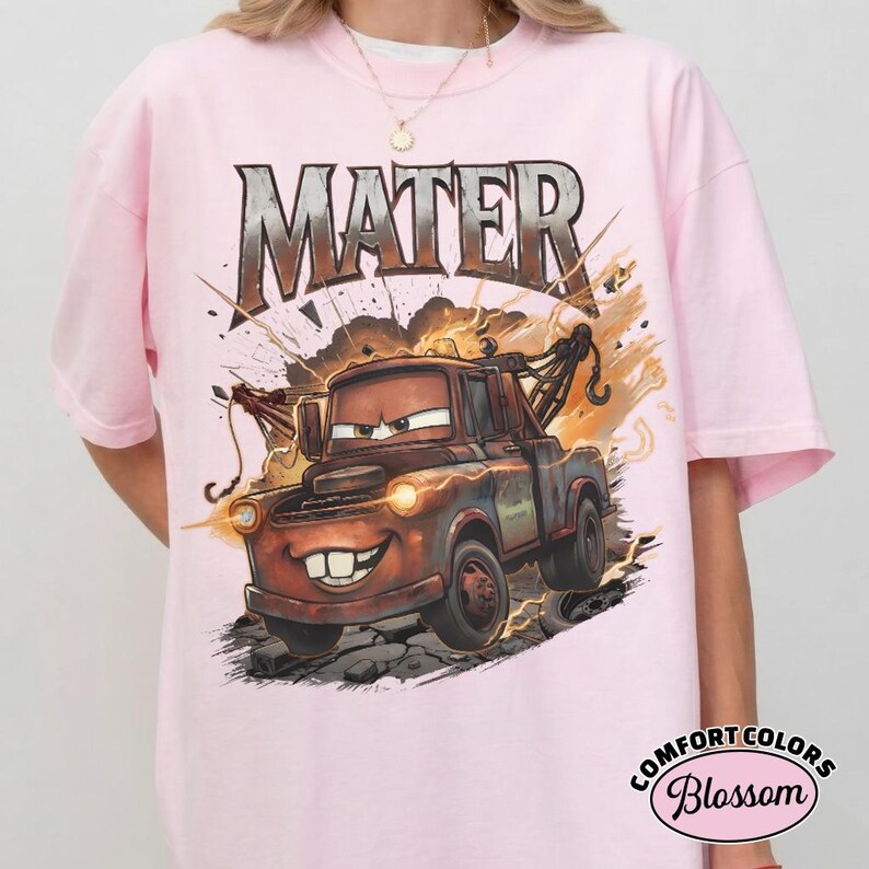 May include: Pale pink t-shirt with a cartoon illustration of Mater from the film Cars. The word "MATER" is printed above the character in a metallic font. The design includes lightning bolts and a distressed background.