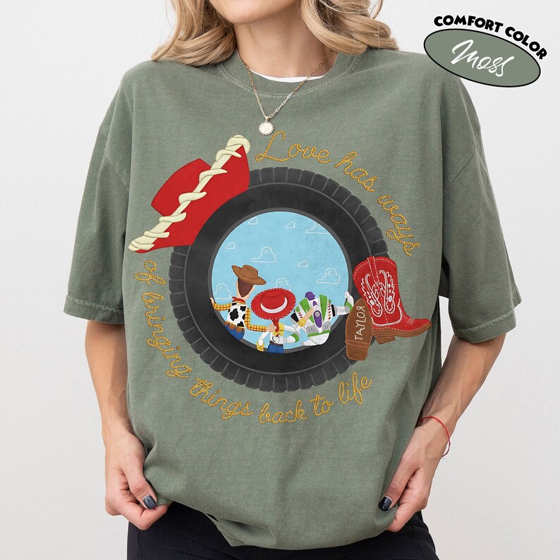 Może przedstawiać: Mchowo-zielony t-shirt z kr&oacute;tkim rękawem z grafiką czarnej opony otaczającej scenę z niebieskim niebem i trzema postaciami z kresk&oacute;wek. Wz&oacute;r zawiera czerwony kowbojski kapelusz, czerwony kowbojski but oraz napis Love has ways of bringing things back.