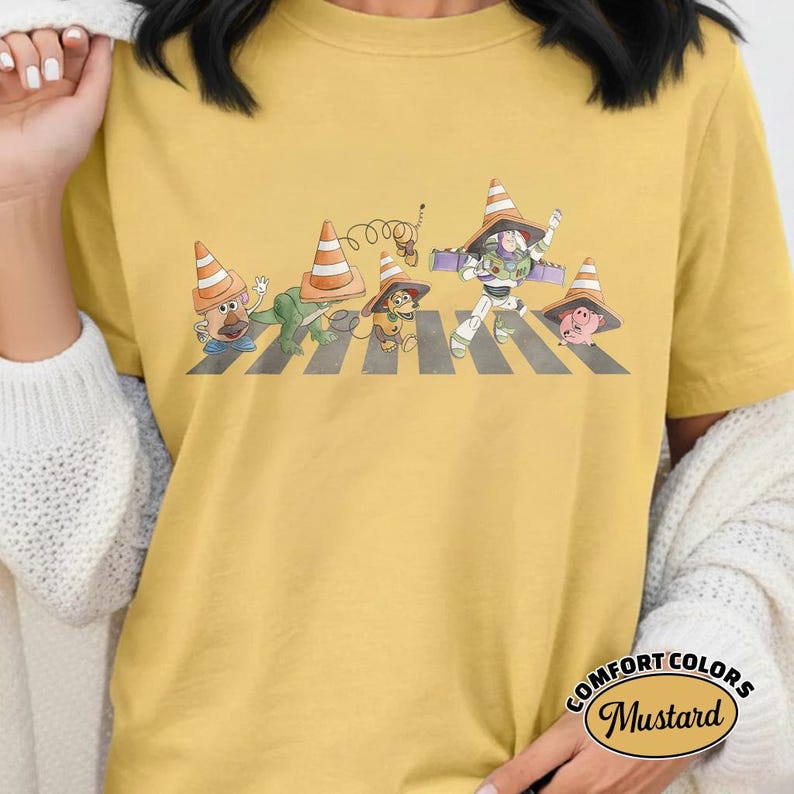 K&ouml;nnte beinhalten: Senffarbenes T-Shirt mit einer Grafik von Toy Story-Charakteren, die einen Zebrastreifen &uuml;berqueren, jeder mit einem Verkehrskegel-Hut. Der Text "COMFORT COLORS Mustard" ist unten rechts aufgedruckt.