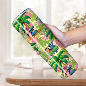 May include: A stainless steel tumbler featuring a tropical design with palm trees, pink flowers, and cartoon characters. The tumbler is cylindrical and has a silver rim. The design includes the characters Stitch and Lilo.