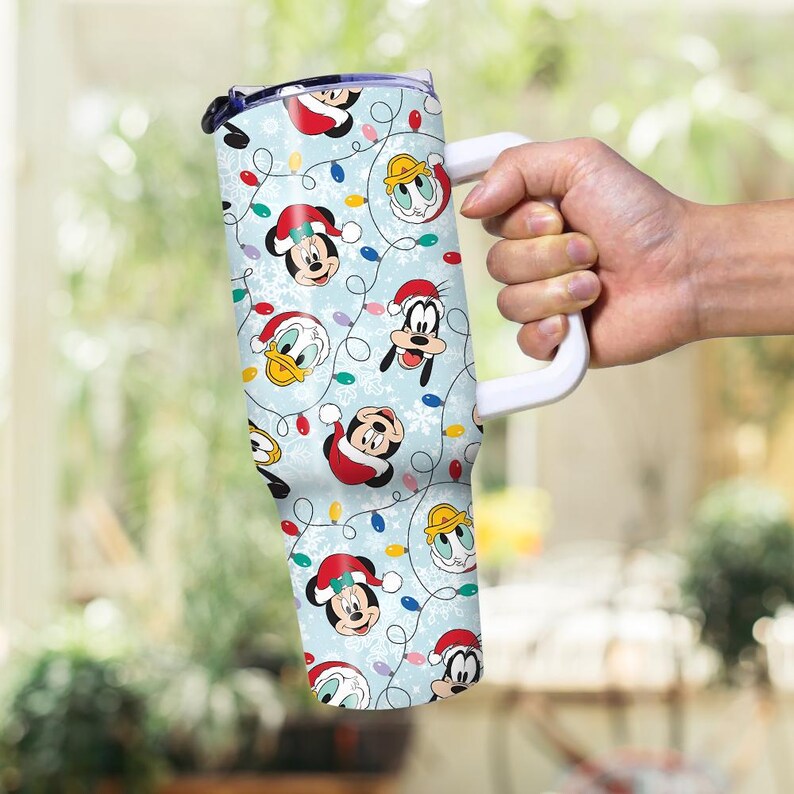 May include: A light blue tumbler featuring a festive pattern of Mickey Mouse, Donald Duck, and Goofy in Santa hats, along with Christmas lights and snowflakes. The tumbler has a white handle and a black lid.