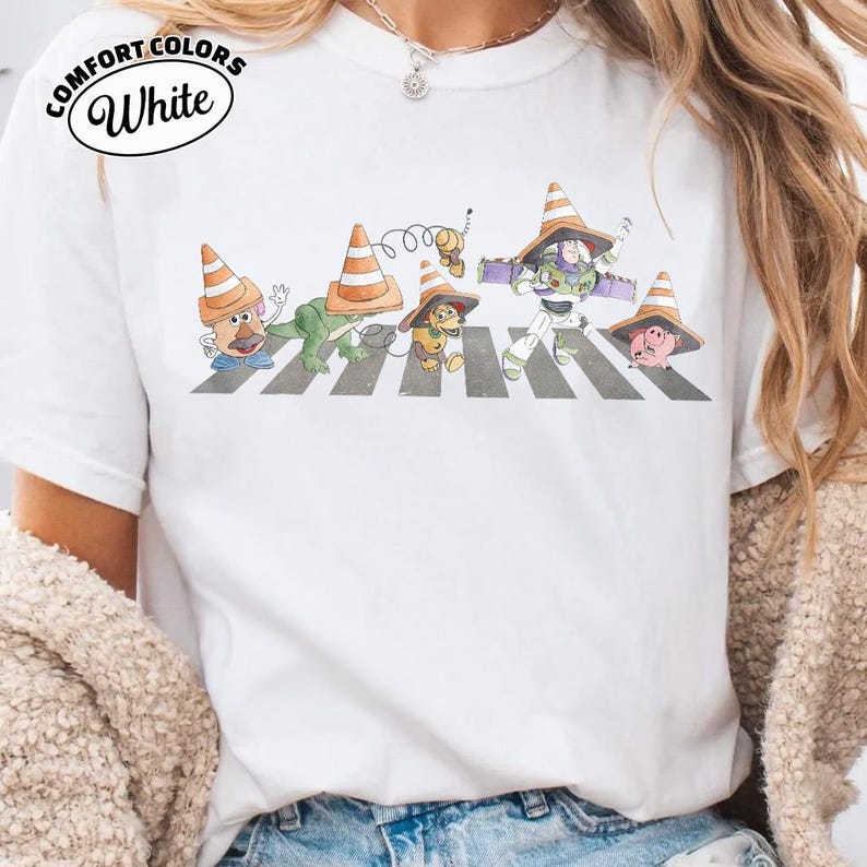 K&ouml;nnte beinhalten: Wei&szlig;es T-Shirt mit einer Grafik von Toy Story-Charakteren, die einen Zebrastreifen &uuml;berqueren, jeder mit einem Verkehrskegel-Hut. Der Text "COMFORT COLORS White" ist oben links aufgedruckt.