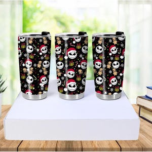 May include: Three stainless steel tumblers with black backgrounds and a repeating pattern of Jack Skellington faces wearing Santa hats, gingerbread men, presents, and snowflakes. The tumblers have black lids and silver bases.